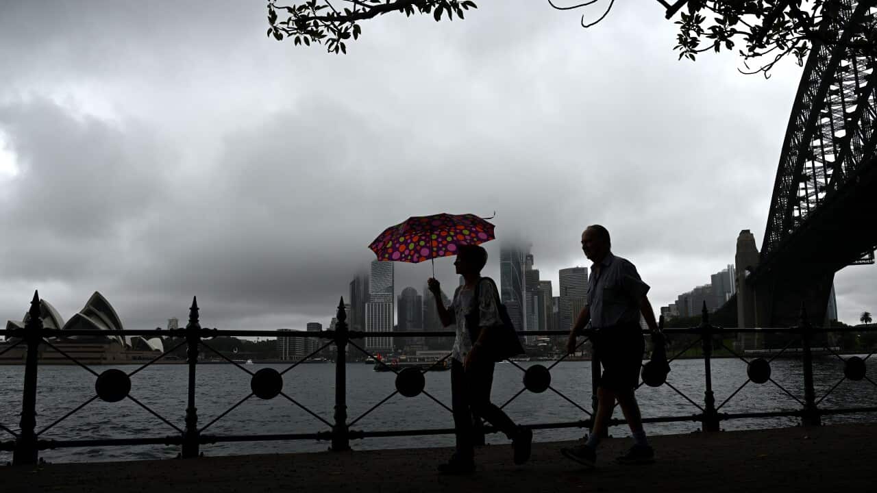Two people walk alongside Sydney harbour, one holding an umbrella, as menacing weather looms.