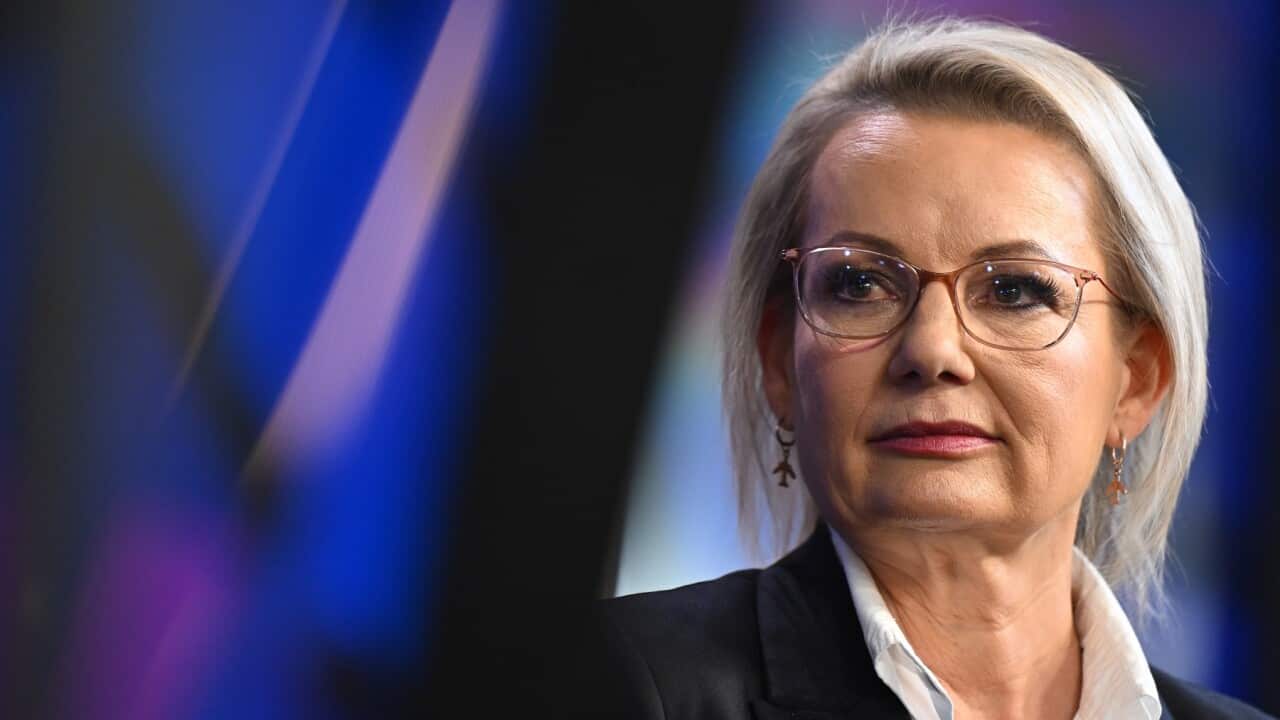 A blonde woman stands in front of a lectern, looking to her left with her lips pursed.