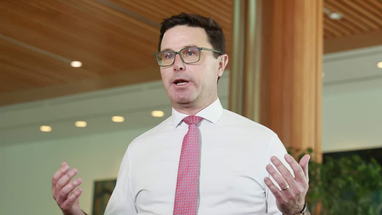 A middle-aged white man wearing a white shirt, red tie and glasses. His hands are slightly raised beside him as he speaks at a press conference in Parliament House in Canberra.