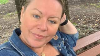 A woman sits on a park bench, her head resting on her hand, with a neutral expression on her face.