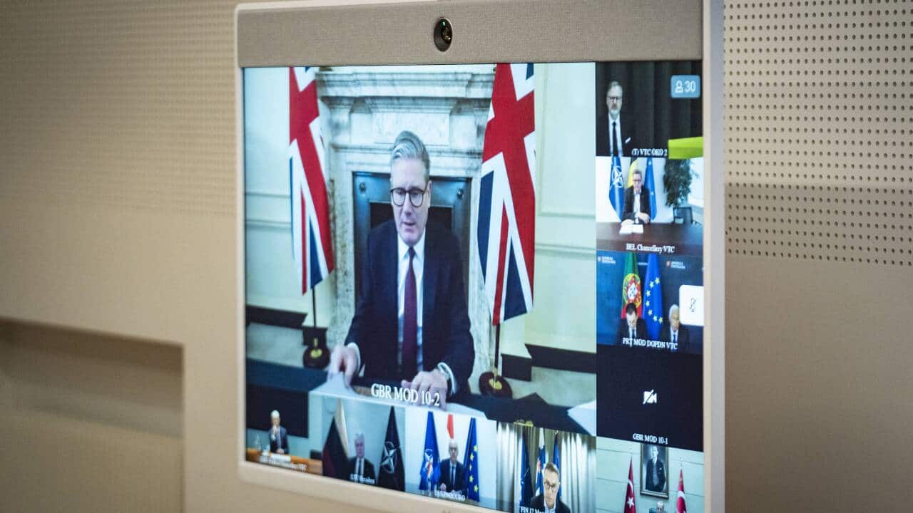 Man in suit and glasses sits at desk with flags on video call with other men.