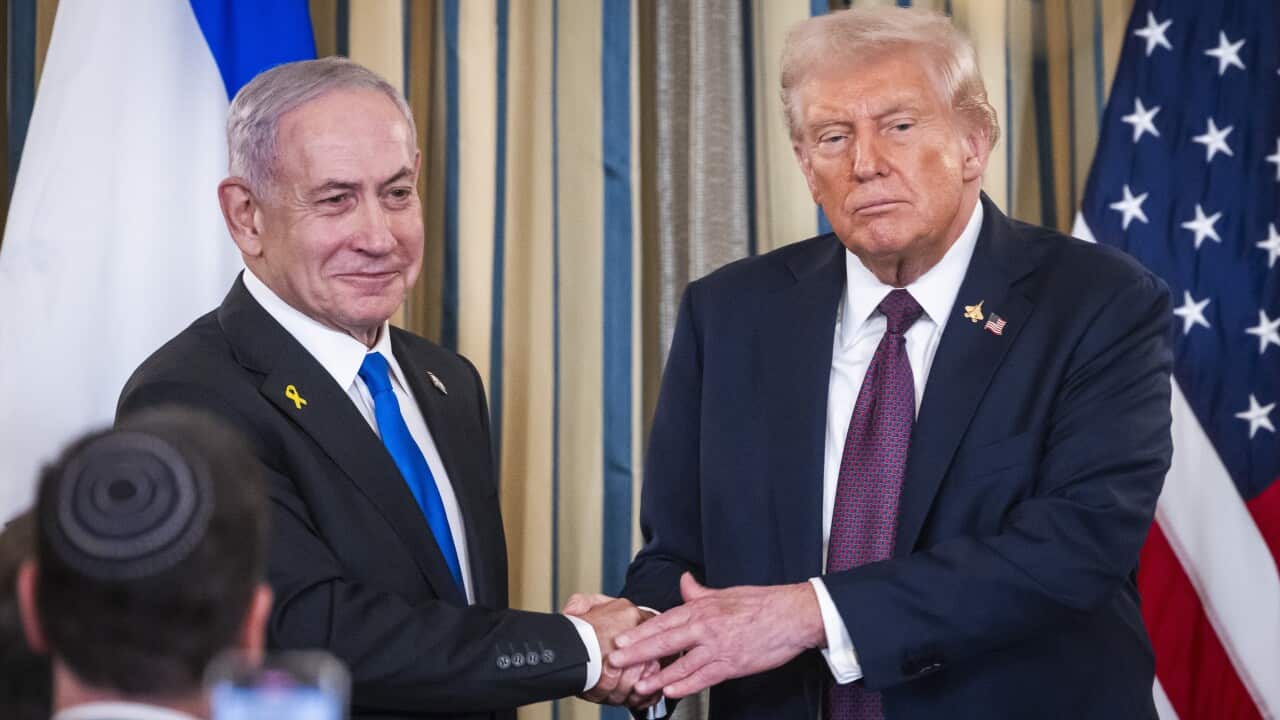 A man in a dark suit shakes hands with another man in a dark suit standing in front of the American flag