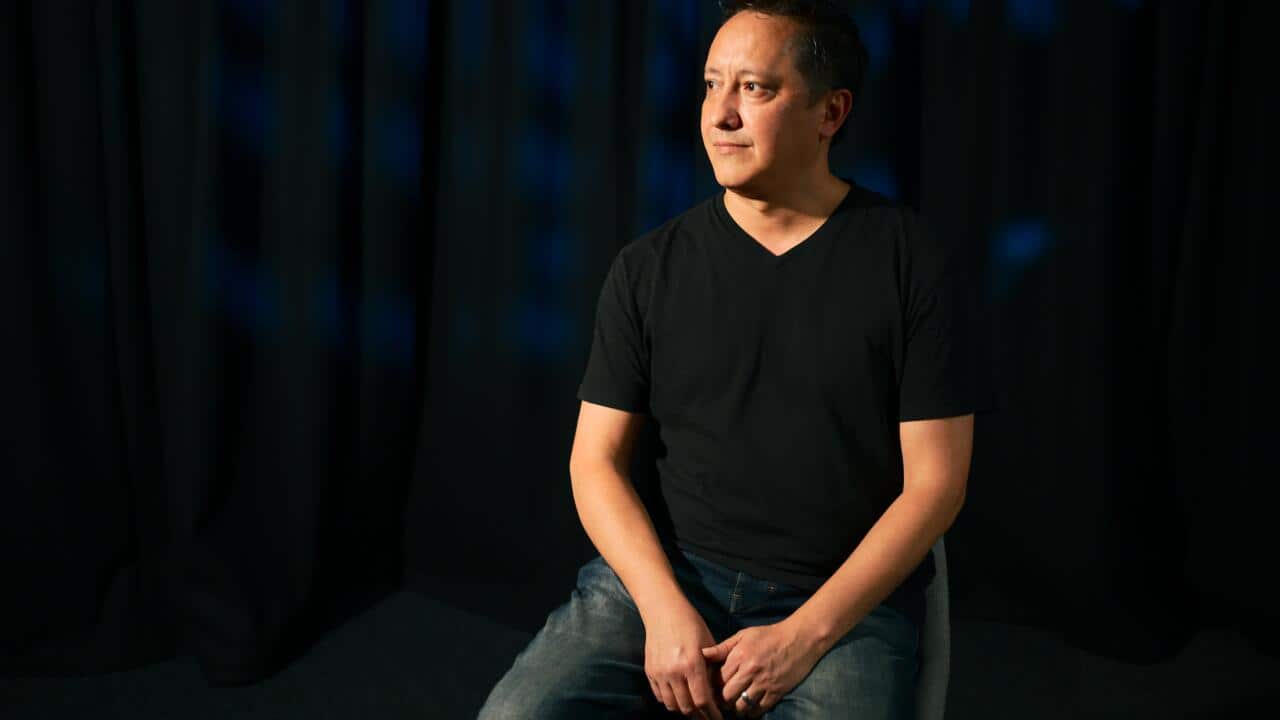 A middle-aged man wearing a black t-shirt and jeans seated in chair. He is looking to his right.