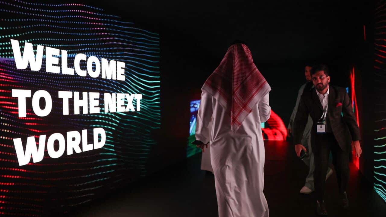 The man in a long white Arabic robe walks past a sign that reads "Welcome to the New World", while a man in formal attire approaches him.