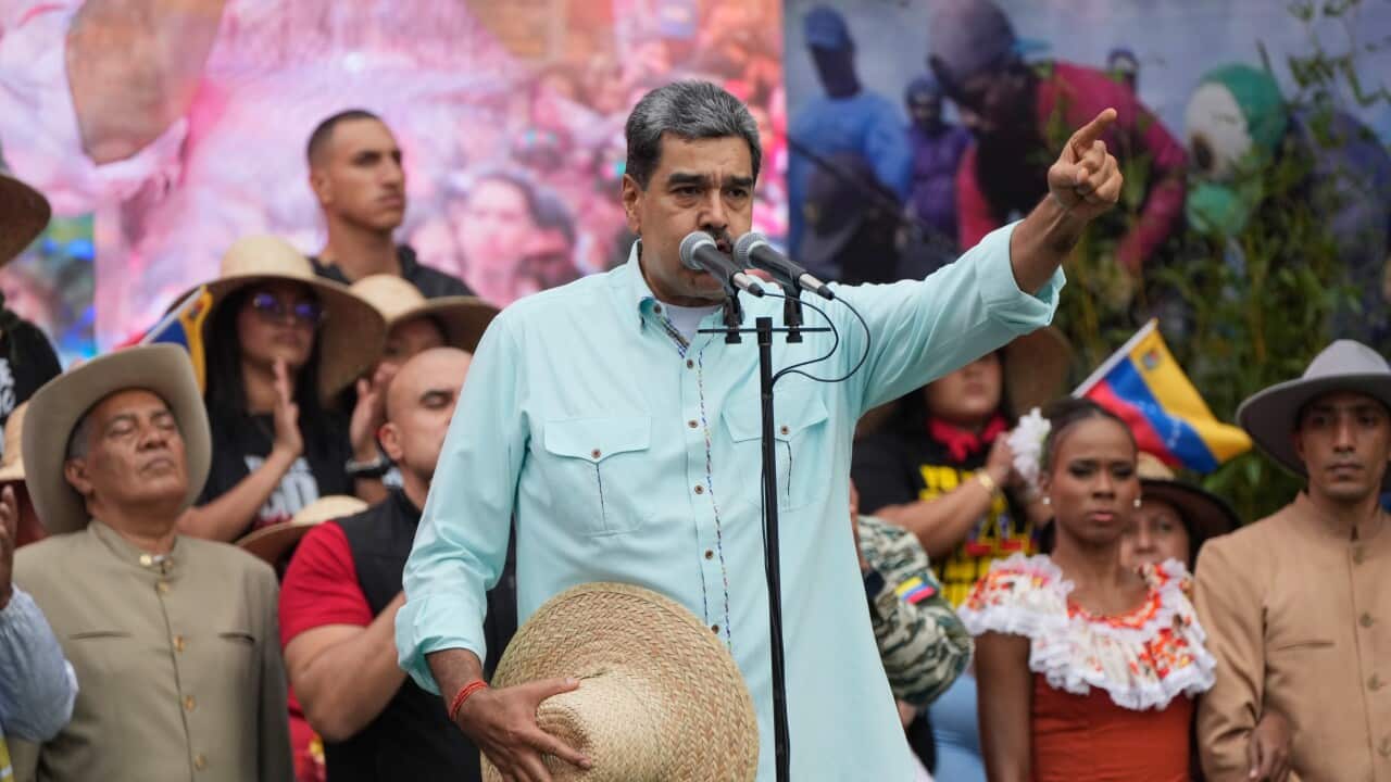 Venezuelan President Nicolás Maduro gestures toward a crowd while speaking at a podium, holding a straw hat in front of supporters dressed in traditional and casual attire.