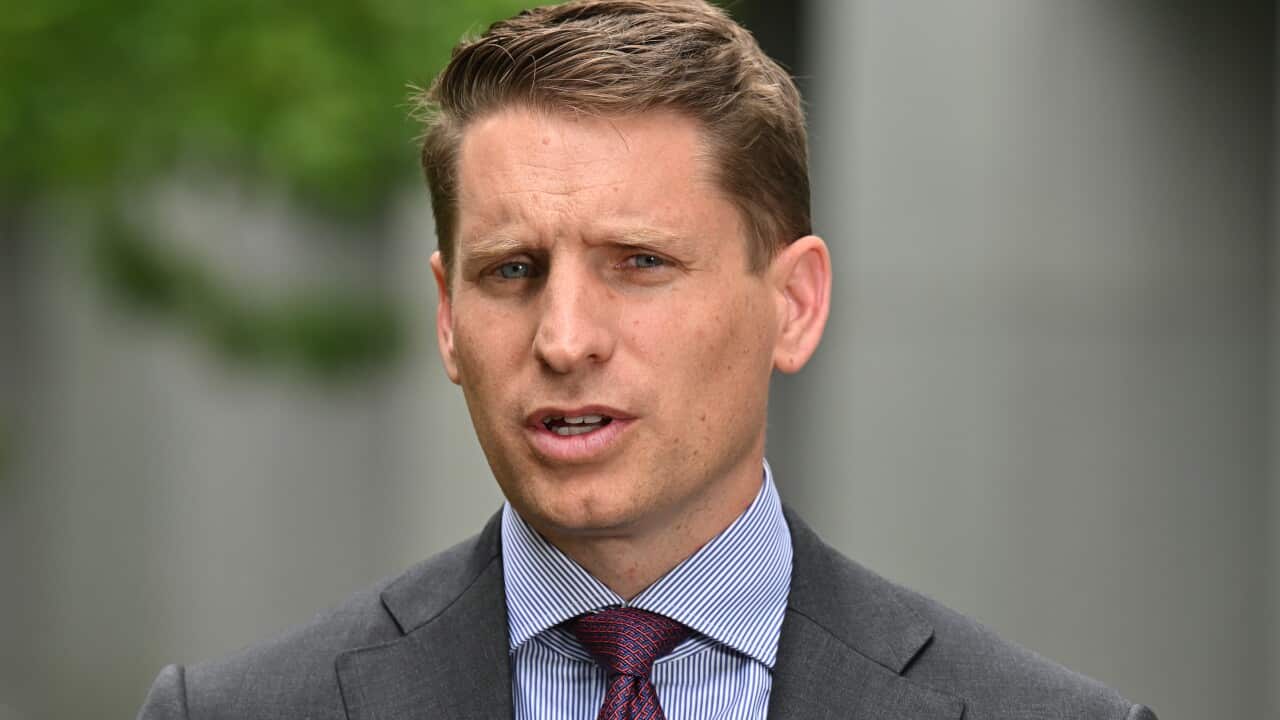 A close-up photograph features a man in a grey suit, striped blue shirt, and a patterned red tie, speaking outdoors with a serious expression. He has short, styled brown hair and appears to be a politician or public figure delivering a statement.
