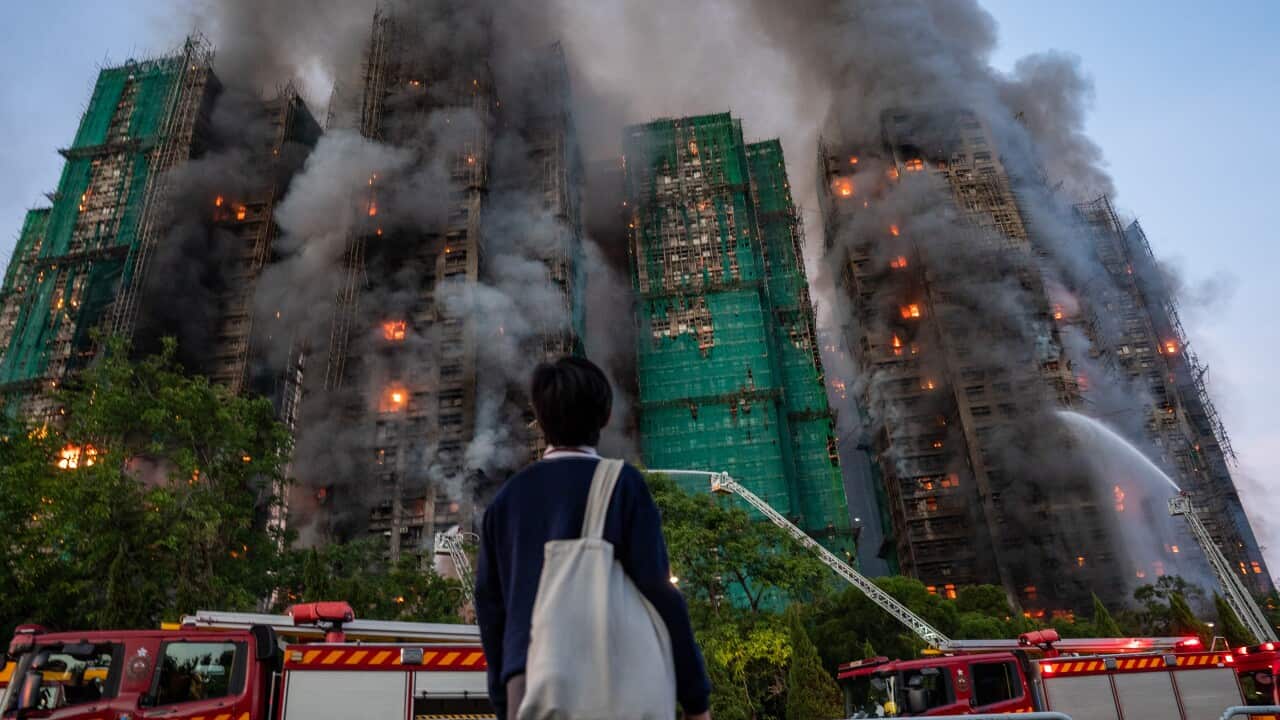 A man wearing a blue jumper and carrying a white tote bag looks on as hoses from firetrucks douse a building complex.