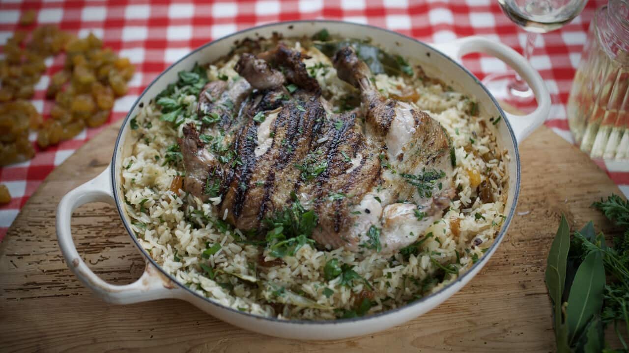 A round white cast iron pan sits on a wooden board, on a red checked tablecloth. It holds a chicken and rice dish, scattered with herbs.