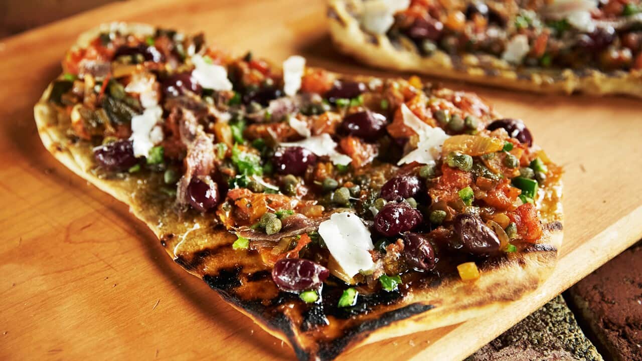 A roughly rectangular pizza with a chunky topping sits on a wooden board. Another pizza xan be seen behind it.