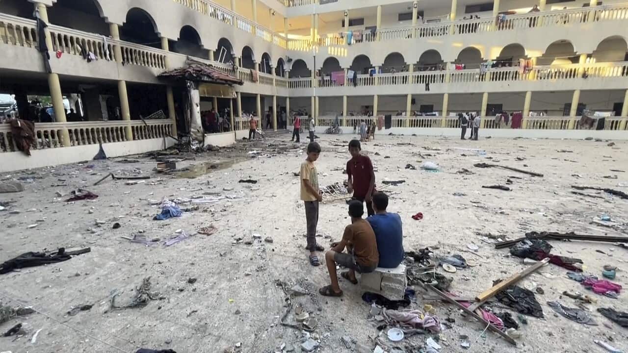 Several young boys sit in a courtyard of a building scattered with debris. Clothing and rugs hang over the banister of the building, parts of which look damaged by explosions.