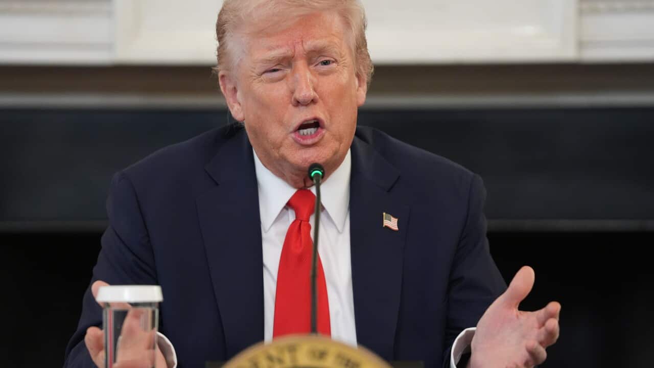 US President Donald Trump, wearing a suit and tie, is speaking in front of a microphone.