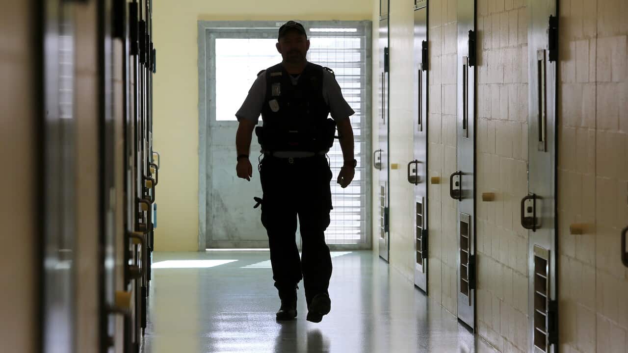 A male prison guard in uniform walks down a corridor which is lined with doors to prison cells.