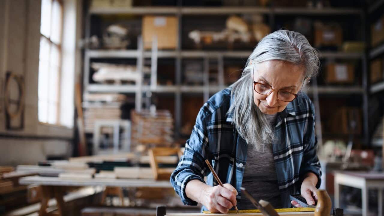 An elderly woman with long gray hair and glasses is focused on her work in a workshop. She's wearing a plaid shirt and is using a small tool, possibly a paintbrush or a carving tool, to work on an object in her hands.