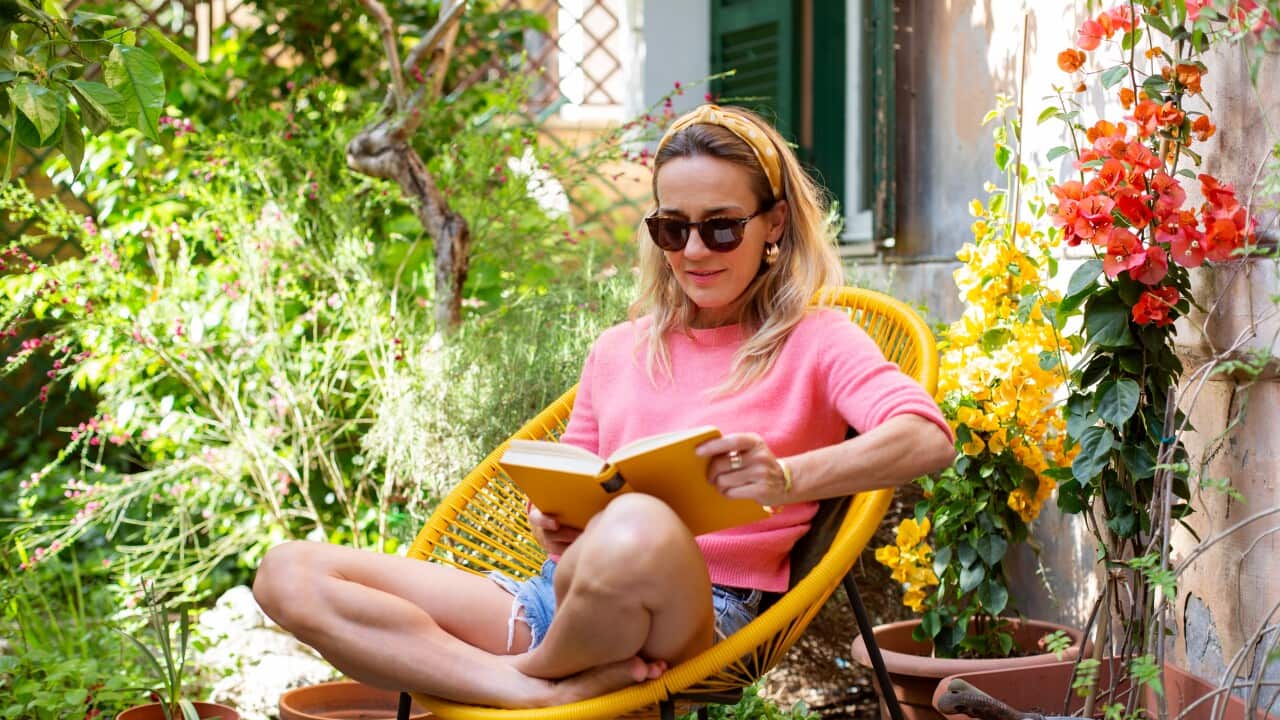 A woman in a pink sweater and sunglasses sits cross-legged in a yellow chair, reading a book in a sunlit garden filled with potted plants and vibrant flowers.