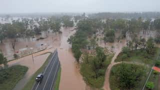 An overhead photo of flooding in the Queensland town of Clermont, taken in January 2026 during Tropical Cyclone Koji. Brown water covers a roadway where a car sits close to the water's edge. Trees stand above the water and, above them, a grey, rainy sky.