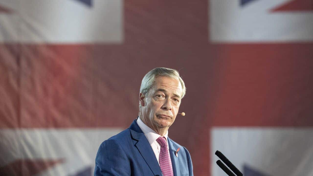 A man wearing a blue blazer, white shirt and red tie, standing in front of a giant British flag.