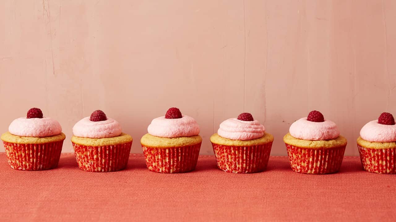 Six cupcakes sit in a row along an orange surface. Each is in a patterned reddish cupcake liner and has a round of pink icing, topped with a single fresh raspberry.
