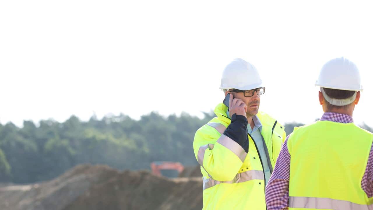 A man wearing a hi-vis jacket speaks into a mobile phone while standing next to another man in a vi-vis vest at construction site.
