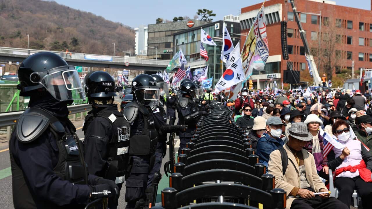 A row of riot police stand in a line in front of a large crowd, some of whom are holding South Korean flags.