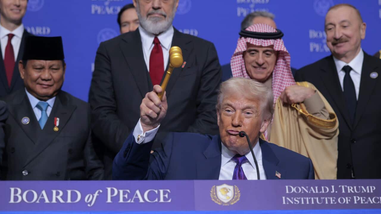 A man in a suit holds up a gold gavel while speaking at a podium labelled “Board of Peace – Donald J. Trump Institute of Peace,” with several formally dressed leaders standing behind him against a blue backdrop.