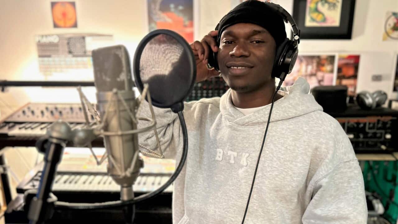 A man in a grey hoodie stands and black beanie at a mic in a recording studio.