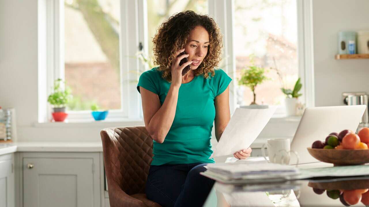 A woman with light brown curly hair speaks on the phone while sitting at a table in a white kitchen. She is holding a piece of paper and has a laptop in front of her