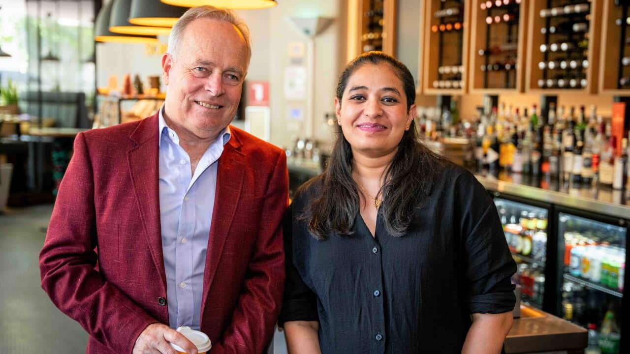 A man in a burgundy jacket stands in a bar next to a woman with long brown hair in a dark blue shirt.