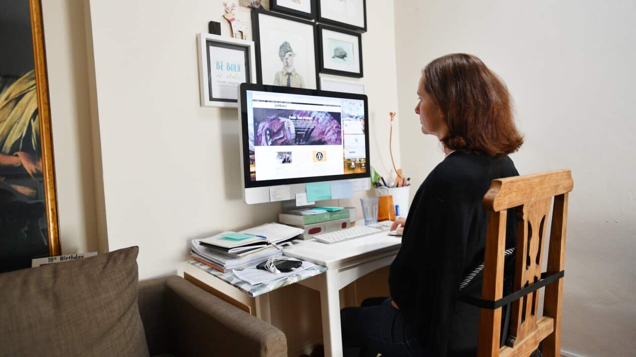 A woman with reddish-brown hair sits at a white desk, looking at a desktop computer monitor. She's wearing a black long-sleeved top and dark pants. The desk has a keyboard, mouse, and various office supplies. Behind her is a light-colored wall with framed pictures. To her left is a brown sofa.