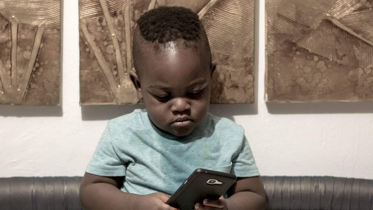 Toddler Holding Mobile Phone While Standing On Bed Against Wall