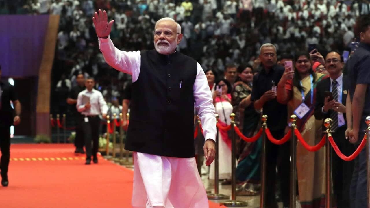 A man in traditional Indian dress raises one hand in the air walking down a red carpet