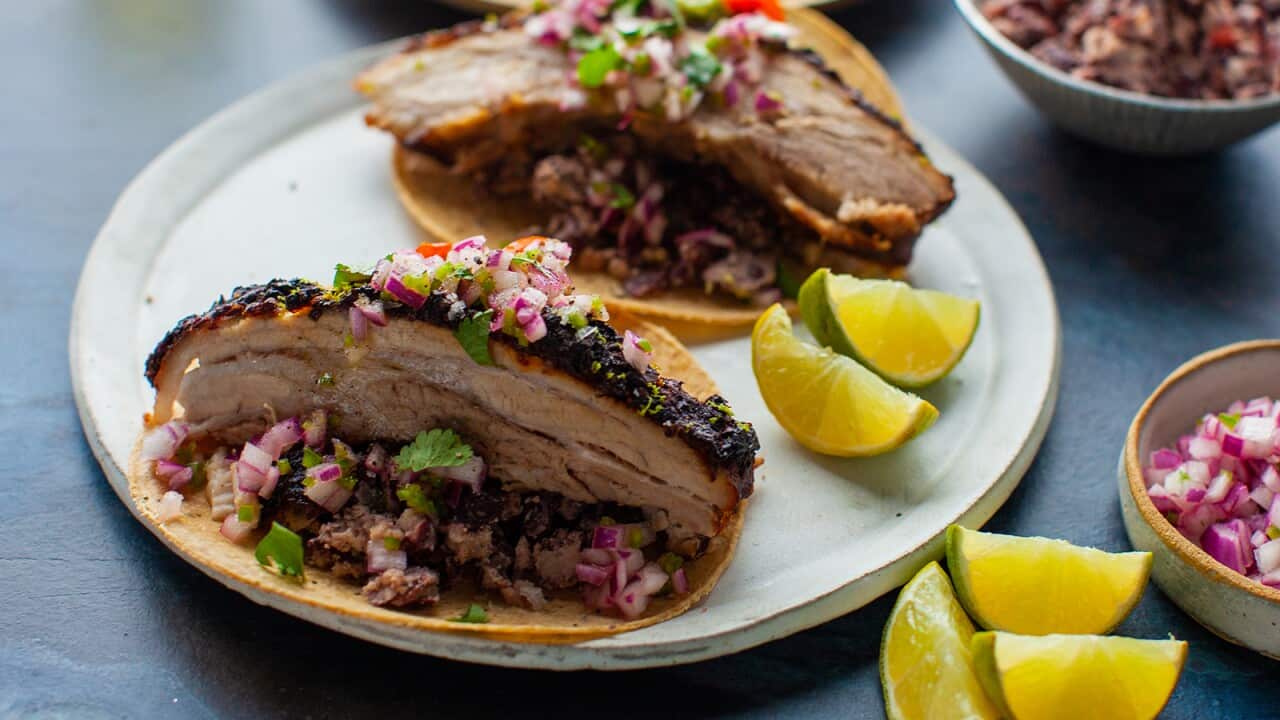 Two tortillas sit on a pale plate, each topped with a generous slice of cooked pork belly, topped with chopped red onion salsa.