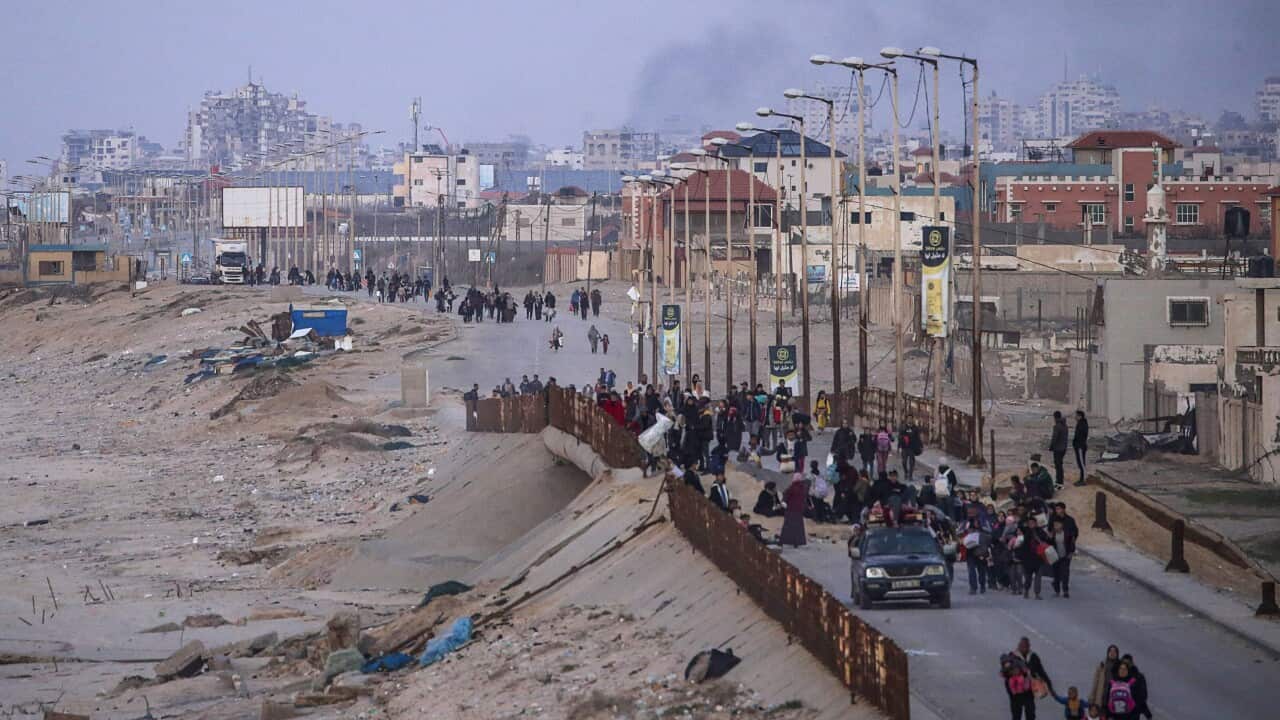 Groups of people walk along a coastal road carrying their belongings. Smoke rises from buildings behind them.