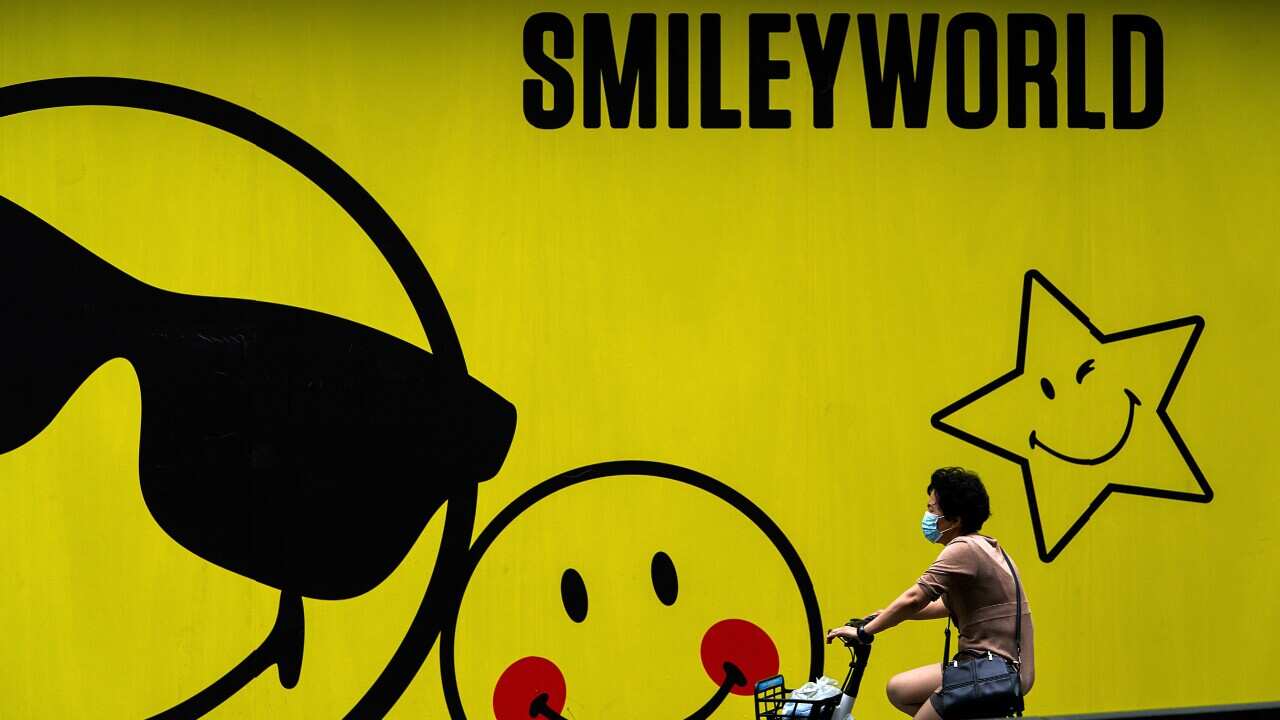 A woman wearing a face mask to help curb the spread of the coronavirus bicycles past a smiley faces billboard outside a shopping mall in Beijing, Monday, July 19, 2021.