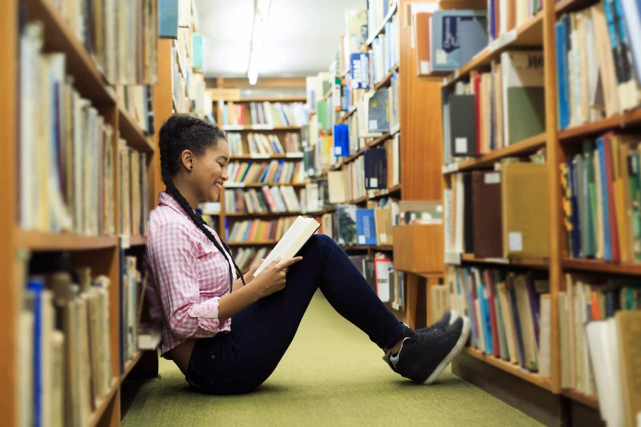 A teenage girl sitting on the floor of a library smiling while reading a book