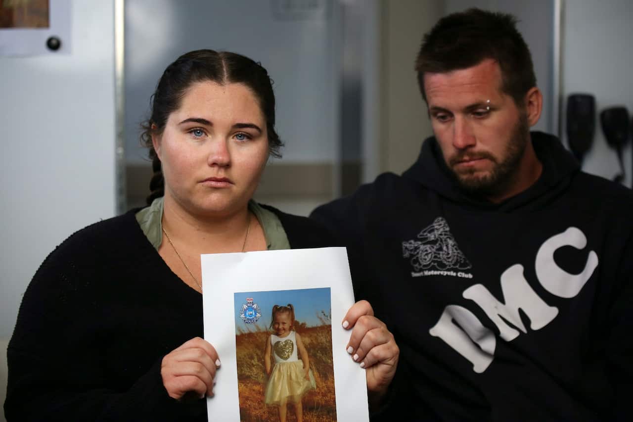 A woman standing inside holds up a picture of a small girl. There is a man standing just behind her