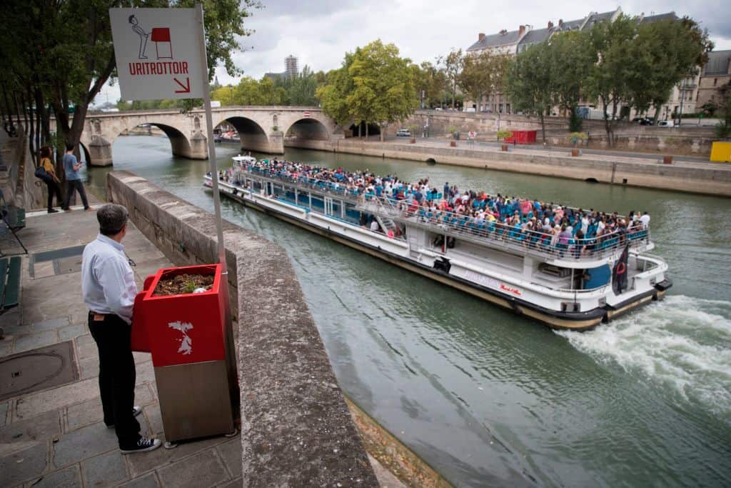 A man stands at a 'uritrottoir' public urinal on August 13, 2018, on the Saint-Louis island in Paris, as a 'bateau mouche' tourist barge cruises past.