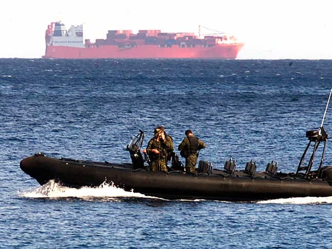 An SAS speed boat travels across the water with a large container vessel in the background.