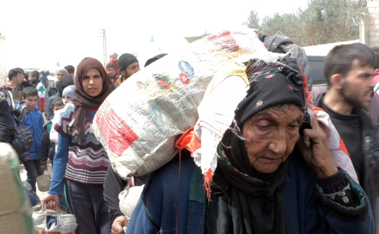 An elderly woman joins the mass evacuation, carrying her possessions on her back. 