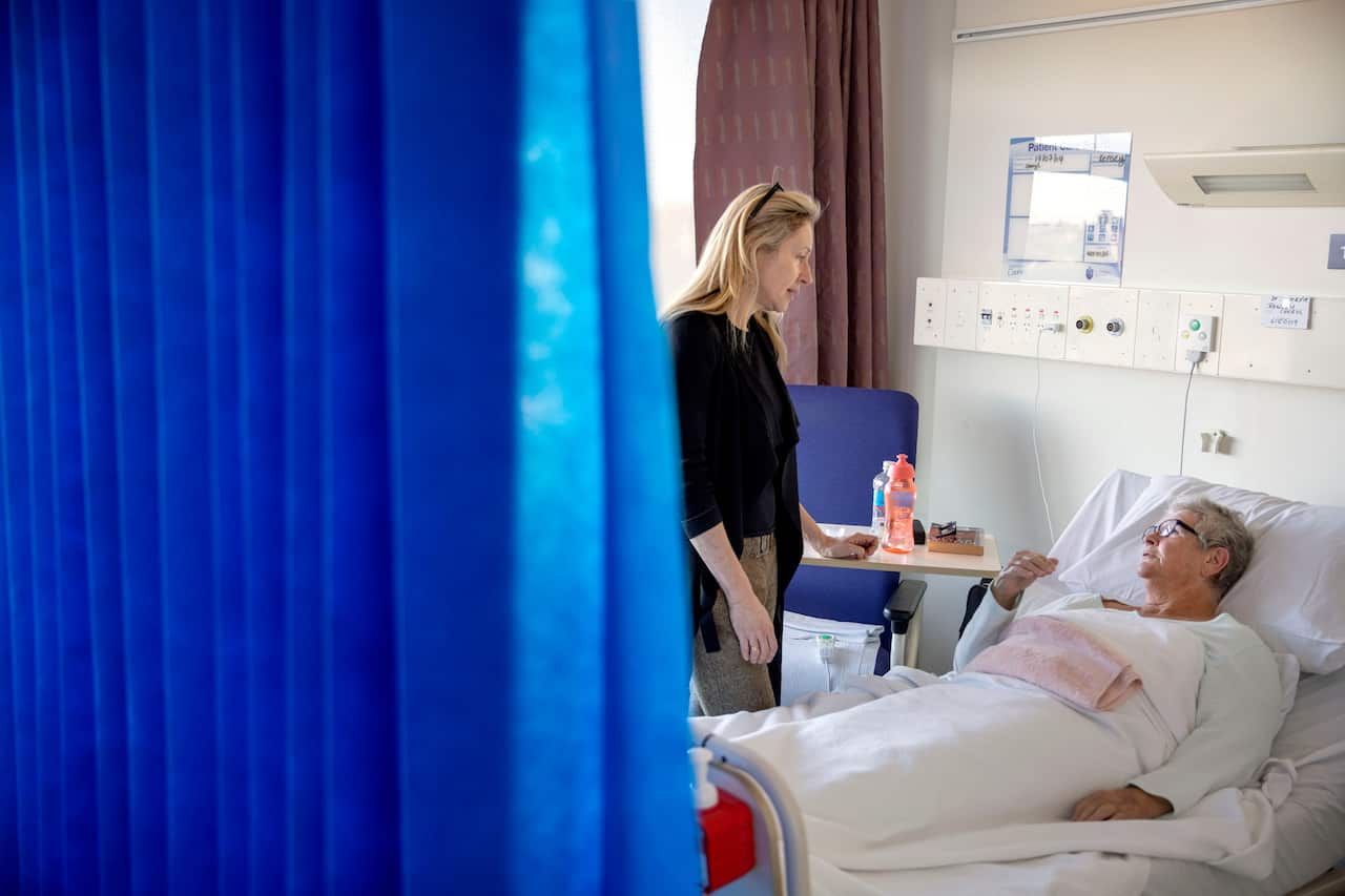 A woman in a black top with blonde hair stands next to a patient lying in a hospital bed. 