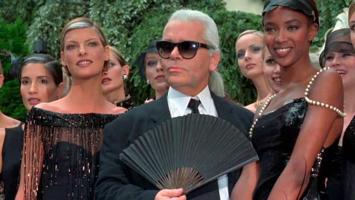 (left to right) Linda Evangelista wearing a black dress, Karl Lagerfeld man wearing a black suit with white shirt and a black tie and Naomi Campbell wearing a black dress. Models stand behind them.
