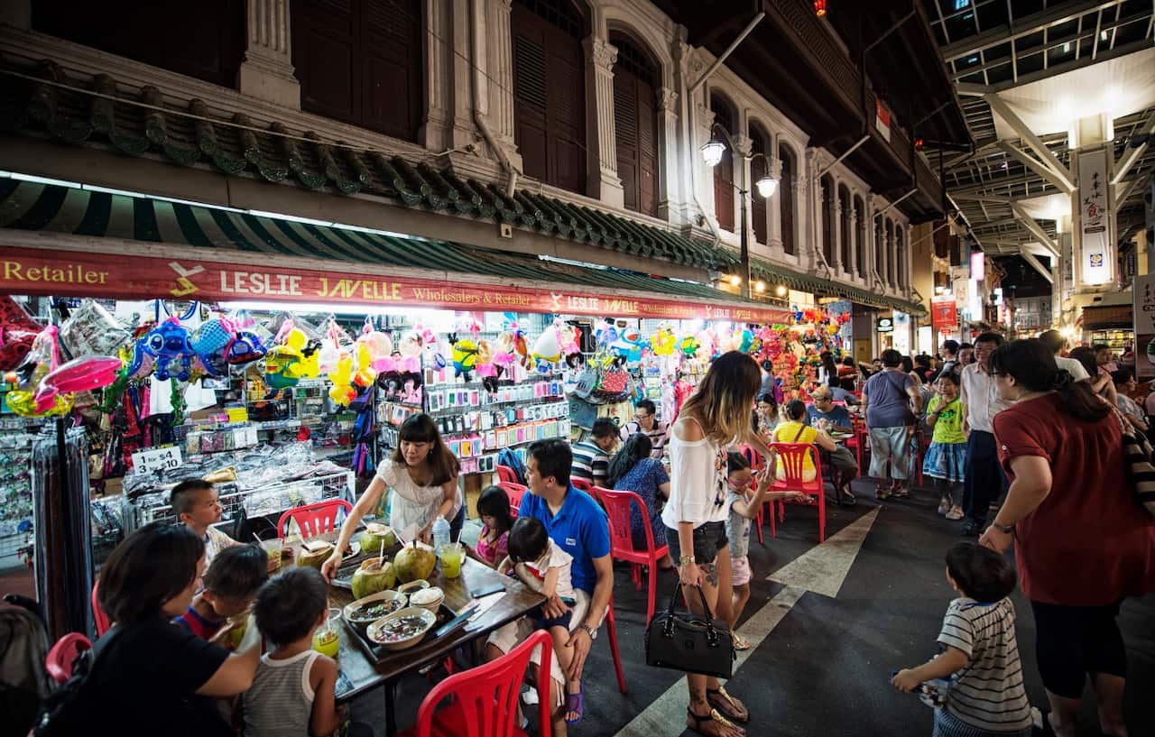A waitress serves diners sitting at a restaurant table as pedestrians walk along Chinatown Food Street in the area of Chinatown in Singapore