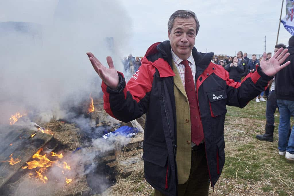 Former UKIP Leader Nigel Farage stands beside the remains of a small boat on top of a bonfire.