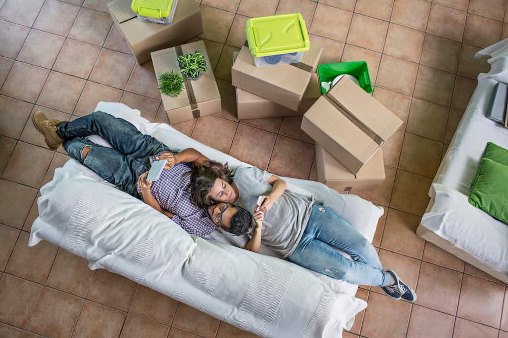 Couple checking their own digital devices on the social networks on the sofa with all the boxes to unpack at the new home.