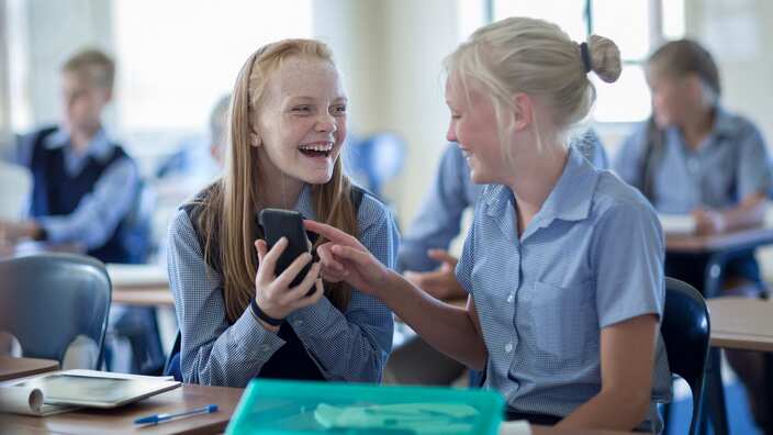 Two happy schoolgirls in classroom with cell phone