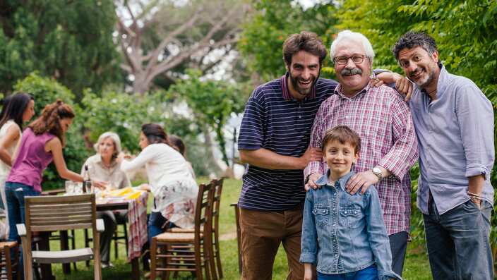 Family at gathering in garden - male family members in foreground, female family members at table in background