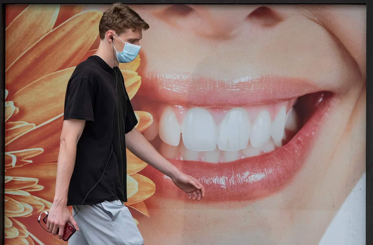 A young man wearing a face mask to curb the spread of COVID-19 walks past a photograph of a woman smiling outside a dental office, in Vancouver, on Monday, August 3, 2020. (Darryl Dyck/The Canadian Press via AP)