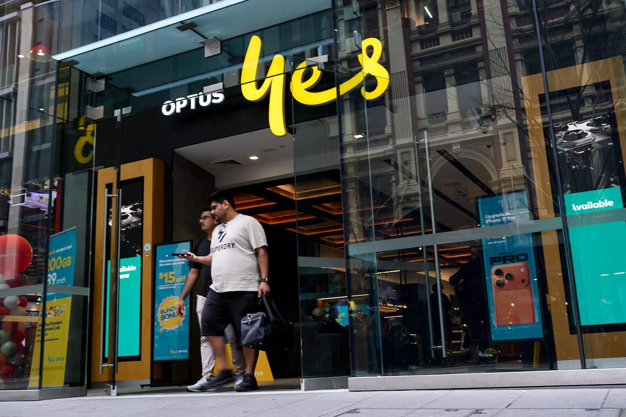 A wide-angle street-level shot shows the entrance to an Optus store with the company's "yes" logo in bright yellow above the door, as two men walk out of the glass entrance.