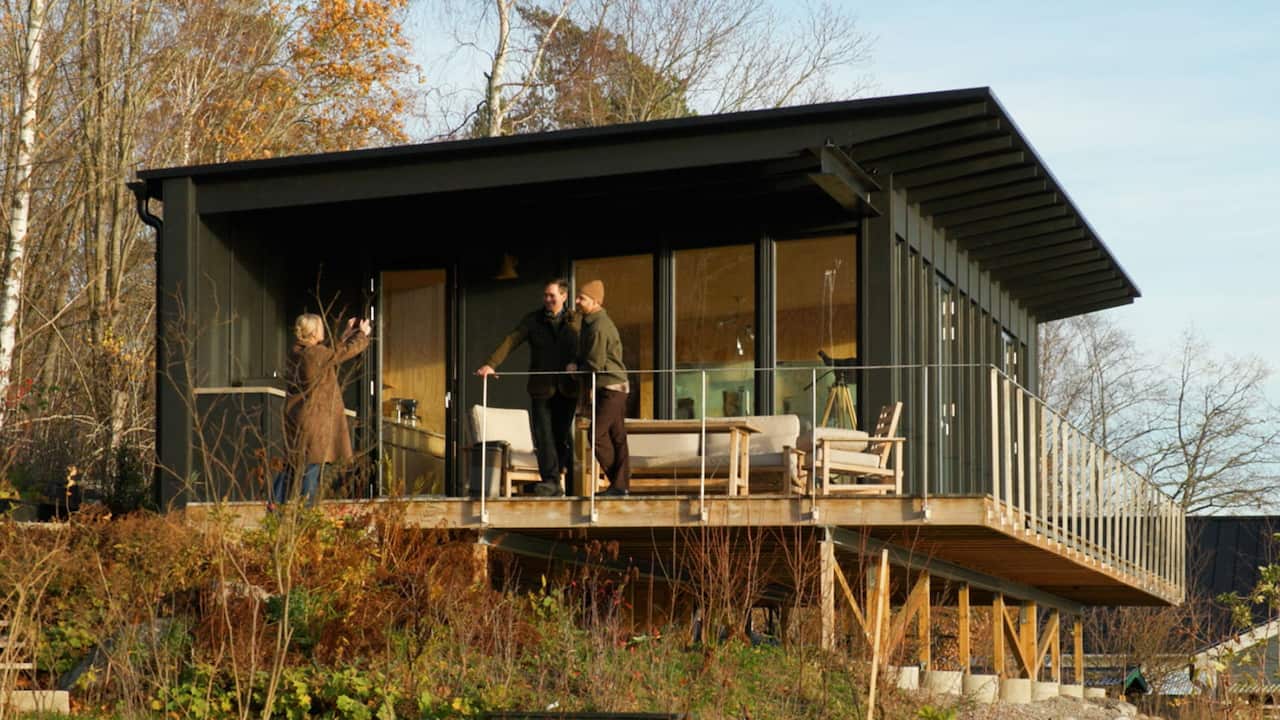 Two men are having their photo taken on a verandah of a wooden house by a woman in a long brown coat