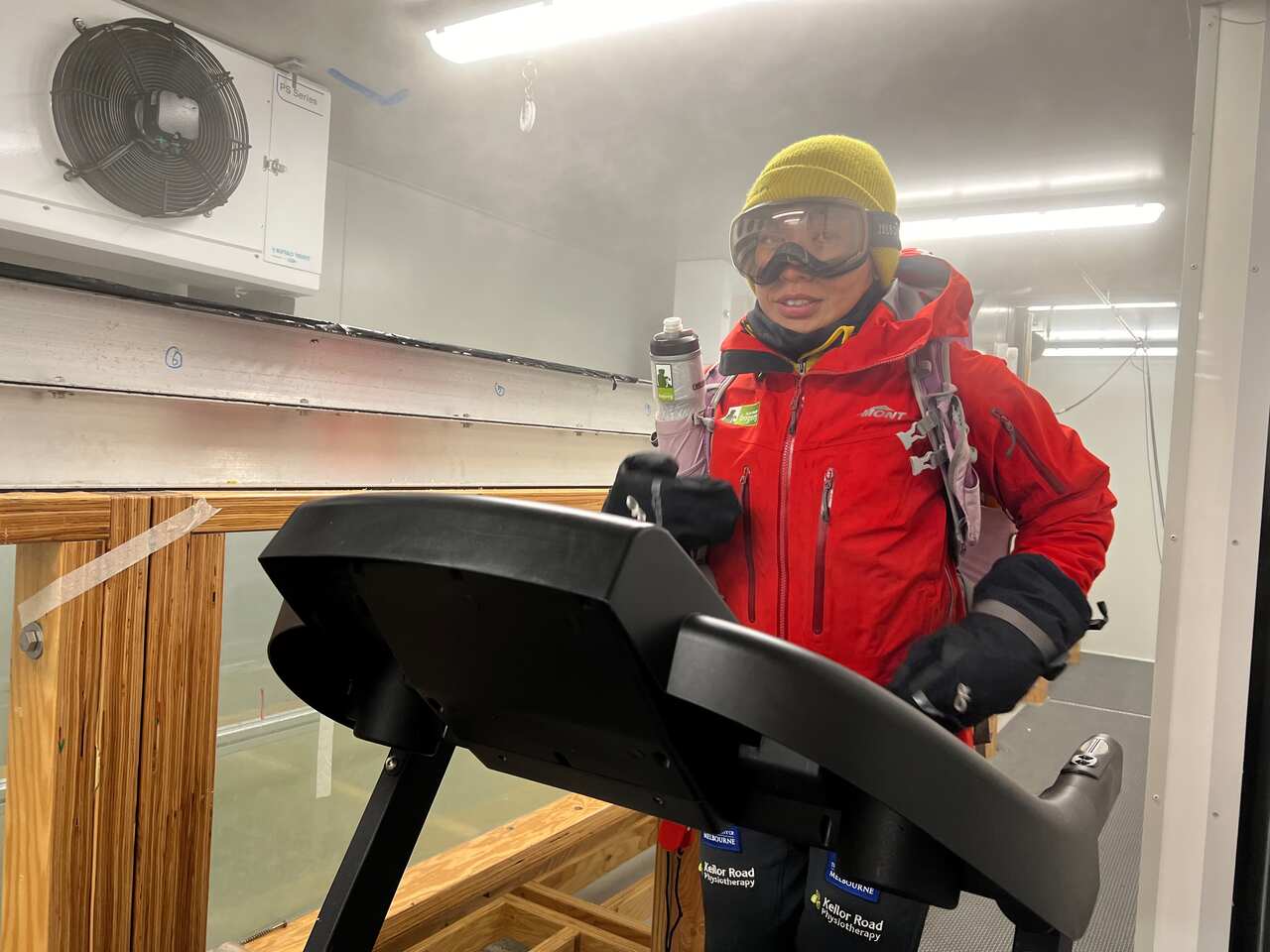 A woman wearing an orange parka, goggles and a yellow beanie runs on a treadmill inside a giant fridge, with cold mist surrounding her.