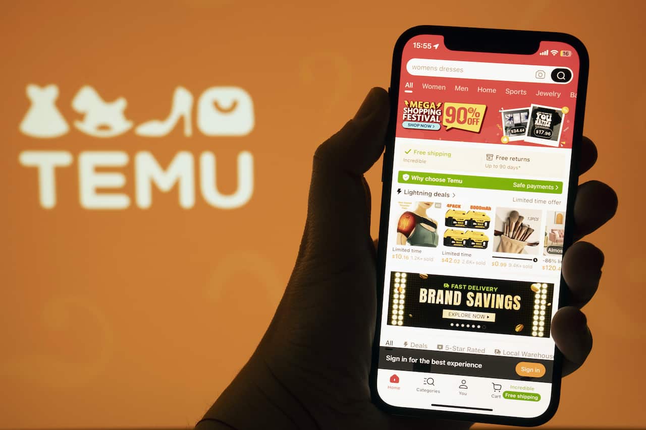 A silhouetted hand holding a phone with the Temu app open, in front of an orange wall that has the Temu logo on it.